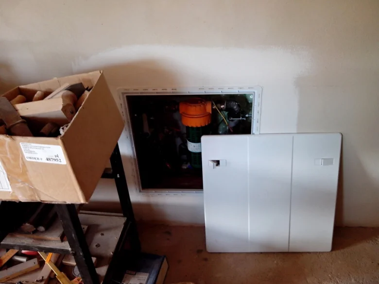 Přístupový panel pro rozvod vody v domě s viditelnými potrubími.