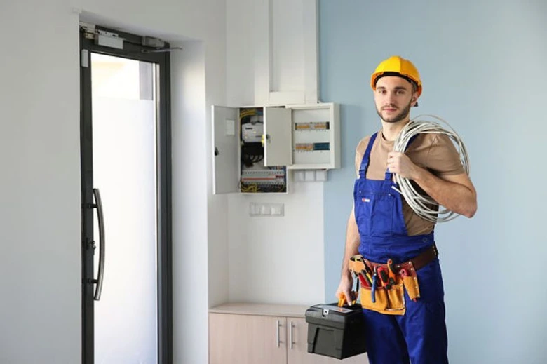 Elektrikář v modrém obleku s nářadím a kabely u rozvaděče.