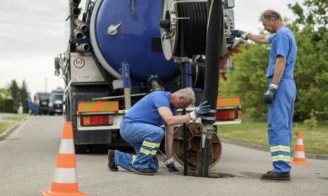 Dva pracovníči čistí septik pomocí velkého nákladního vozu.