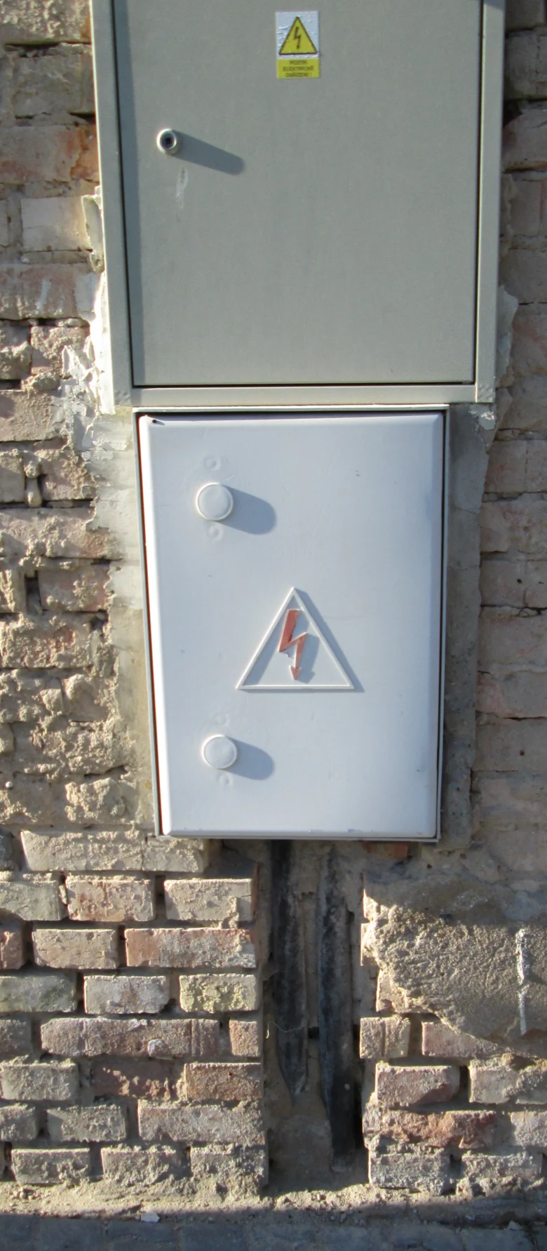 Dvě šedé elektrické skříně na cihlové zdi s varovnými symboly.